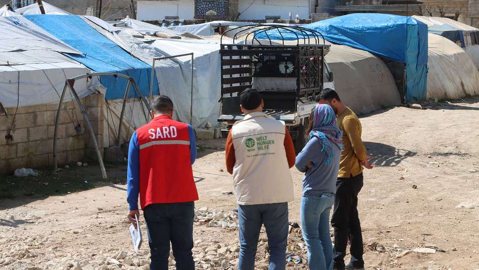 Vier Personen, zwei von ihnen in Uniformen von Hilfsorganisationen, in einem Flüchtlingslager in Syrien. Die Umgebung besteht aus provisorischen Unterkünften, die mit blauen und weißen Planen bedeckt sind.